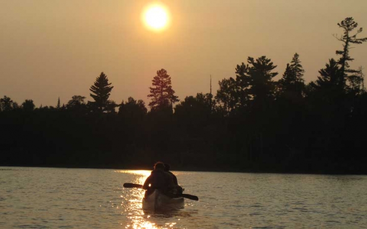 the silhouette of a canoe appears on calm water, with the sun just above the tree-line in the background.